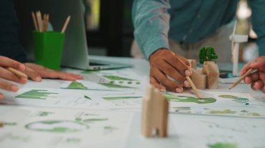 Dinamik bir çalışma alanında, sürdürülebilir tasarım kavramlarını analiz eden ve çizen, takım çalışmalarını ve çevre dostu stratejileri vurgulayan çeşitli bireylerin elleri sergileniyor. SACTR