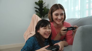 Annem dizüstü bilgisayar kullanarak kanepeye uzanırken, mutlu kız oyun oynuyor ve oturuyor. Beyaz bir ebeveyn ve çekici bir kız, elinde joystick ile birlikte vakit geçirir ve maç kazanmaya odaklanırlar. Pedagoji.