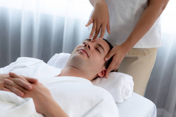 Кавказский мужчина наслаждается расслабляющим анти-стрессовым массажем головы и расслабляющим отдыхом кожи лица в дневном спа современном светлом окружении на роскошном курорте или в спа-салоне отеля. Quiescent