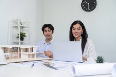 Profesyonel Asyalı erkek mimar ev modelinin uzunluğunu ölçmek için cetvel kullanırken, beyaz tenli genç meslektaşı ev modeliyle toplantı masasındaki verileri analiz etmek için dizüstü bilgisayar kullanıyor. Tertemiz..
