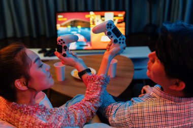 joystickli elleri çapraz oynayan bir çift arka tarafta TV ekranında dövüş oyunu oynuyor, modern rahat evinde neon ışıklı oturma odasında kanepesinde oturuyor. İnfobahn.