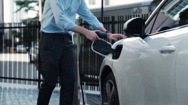 İlerici işadamı, şehir merkezinde dolaşmadan önce şarj cihazından elektrikli arabasına şarj prizi takıyor. Çevre dostu şarj edilebilir araba, sürdürülebilir ve temiz enerjiyle çalışıyor..