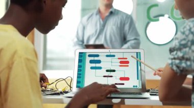 Back view of diverse student coding system while girl pointing at screen. Smart teacher standing while explain about code or prompt while sitting at table with electronic tool placed. Edification