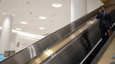 Kafkas proje yöneticisi yürüyen merdiven kullanırken telefonla konuşuyor. Beyaz tenli yönetici, pazarlama fikri ya da planı rapor etmek ve tartışmak için iş ekibiyle konuşuyor. Yan görüş. Sevinçli..