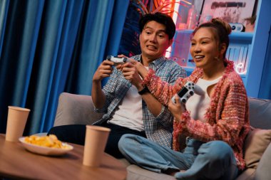 Neşeli, gülümseyen bir çiftin koltuğunda oturup oyun oynuyorlar. Oyun konsolu, hazır aperatifler ve içecekler, modern neon ışıklı, rahat oturma odasında eğlenceli bir rekabet seviyesinde. İnfobahn.