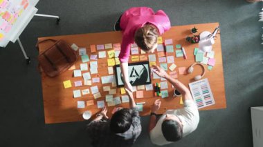 Proje yöneticisinin üst görüntüsü toplantı masasındaki iş adamıyla el sıkışıyor yapışkan notlar ve tablet sergilenen logo tasarımı ile. İş ekibi birlikte çalışıyor ve fikir hakkında konuşuyor. Sempozyum.