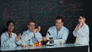 Çok kültürlü bir öğrenci mikroskop altında incelerken güzel bir kız da numuneyi inceliyor. BTMM sınıfında karatahta bilim teorisi deneyi yapan liseli çocuklara bakan öğretmen. Öğretim