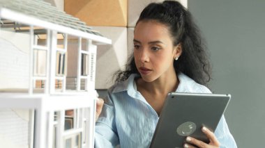 Genç, güzel İspanyol mimar mühendis modern ofisteki ev modeli yapısını kontrol ederken tablet kullanarak bina inşaatını çiziyor. Yaratıcı iş inşaat konsepti. Yönlendirici.