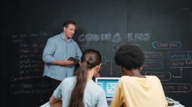 Profesyonel öğretmen tableti tutarken, karatahtadaki programlama sistemini açıklarken, karışık ırklı bir genç BTMM sınıfında dizüstü bilgisayarda kodlama programı ile çalışıyor. Öğretim