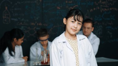 Çeşitli gruplar deney yaparken sevimli Asyalı bilim adamları kameraya gülümsüyor. Laboratuvarda laboratuvar önlüğü giyerken yazılı kimyasal teorisiyle tahtada duran mutlu genç kız öğrenci. Öğretim.