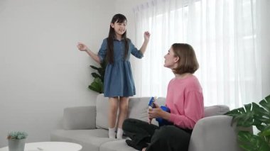 Annem gitar çalıp kıza bakarken yetenekli kız kanepede dans edip zıplıyor. Beyaz anne, çocuk oturma odasında dururken akustik müzik çalar. Aile eğlence konsepti. Pedagoji.