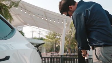 Genç adam, EV şarj aletini şehrin alışveriş merkezinin otoparkındaki şarj istasyonundan elektrik aküsünü şarj etmek için kullandı. Sürdürülebilir şehirde EV aracı tarafından şarj edilebilir enerji olarak kullanıldı.