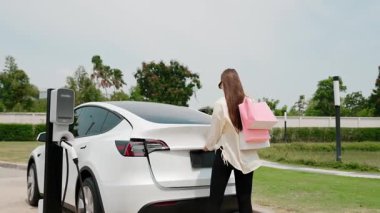 Genç kadın otoparktaki şarj istasyonundan EV araba aküsünü alışveriş çantasıyla şarj ediyor. Modern kadın, şehir içi seyahat tarzında çevre dostu elektrikli araçla alışverişe gider. Yararlı