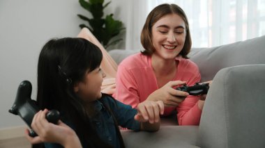 Annem dizüstü bilgisayar kullanarak kanepeye uzanırken, mutlu kız oyun oynuyor ve oturuyor. Beyaz bir ebeveyn ve çekici bir kız, elinde joystick ile birlikte vakit geçirir ve maç kazanmaya odaklanırlar. Pedagoji.