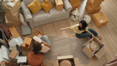 Oturma odasındaki mobilyaların taşınmasına yardım ederken yerde oturan mutlu oda arkadaşı. Yeni eve taşınmak için birlikte çalışan Asyalı lezbiyen aşığının havadan görünüşü. Yer değiştirme konsepti. Myrmidon.