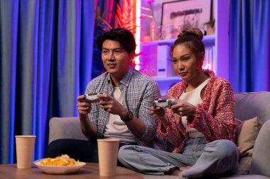 Neşeli, gülümseyen bir çiftin koltuğunda oturup oyun oynuyorlar. Oyun konsolu, hazır aperatifler ve içecekler, modern neon ışıklı, rahat oturma odasında eğlenceli bir rekabet seviyesinde. İnfobahn.