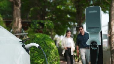 Yavaş çekim çift, ilkbaharda açık yeşil şehir kafesindeki şarj istasyonundan elektrikli araba aküsünü şarj ediyor. Sürdürülebilir çevre dostu seyahat yaşam tarzı için şarj edilebilir EV araba