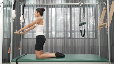 Kadın spor salonunda pilates reformatörüyle çekirdek güçlendirme egzersizi yapıyor. Çekici bayan pilates eğitmeni diz çökmüş vaziyette Cadillac 'ta omuz hareketliliğini arttırmak için bekliyor. Alışkanlık.