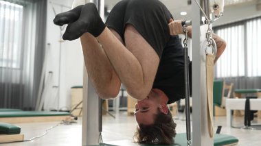 Pilates Cadillac 'ı eğitmen rehberliğinde ters çevirme egzersizi yapan bir müşteri. Çekici bir adam, vücudunun üst kısımlarını güçlendirmek için barda sallanıyor. Çekirdek ve istikrar artıyor. Alışkanlık.