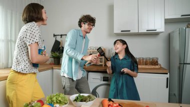 Beyaz yetenekli baba, anne ve Asyalı kız birlikte dans ederken kahvaltı hazırlıyorlar. Modern mutfakta sebze hazırlarken ukulele çalan yetenekli bir anne. Sağlıklı gıda konsepti. Pedagoji.