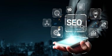 Bir işadamı, arama motoru optimizasyonunu geliştirmek ve modern bir şehirde çevrimiçi görünürlüğü sağlamak için SEO 'yu temsil eden görsel simgeler içeren bir akıllı telefon sergiliyor. XDMCP