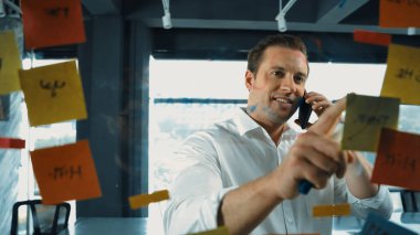 Profesyonel işadamı renkli yapışkan notlara yazılmış pazarlama stratejisi fikrine bakarken müşterilerine telefon ediyor. Proje yöneticisi duvardaki telefon analiz verileriyle konuşuyor. Yönlendirici.