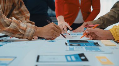 Çeşitli bir ekip renkli çizimler ve araçlarla dolu bir masanın etrafında toplanıyor, kullanıcı arayüzü tasarımı ile meşgul. Atmosfer işbirlikçi ve yenilikçi. SACTR