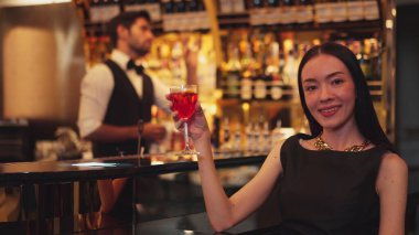 Güzel bir konuk, gece kulübünün mükemmel atmosferinde barmenlerin arka planında çiçeklerle süslenmiş karışık votka Cosmopolitan kırmızı kokteyli ile lüks bir gişede içki içerken kameraya bakıyor. Vinosity.