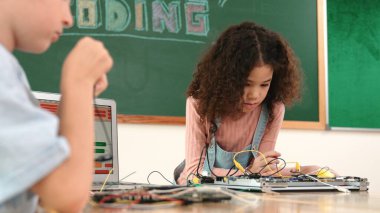 Kız tornavida kullanarak elektronik tahtayı tamir ederken ayakta duruyor. Amerikalı öğrenci ve beyaz çocuk elektrik sistemini denetlemek için birlikte çalışıyorlar. Gemide çalışan meraklı çocuklar. Pedagoji.