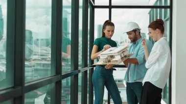 Proje yöneticisi ev modelini elinde tutarken, mimar mühendis takımı da ev tasarımı veya bina inşaatı hakkında fikir alışverişinde bulunuyor. Şehrin penceresinin yanında iç mimar duruyor. İzleme