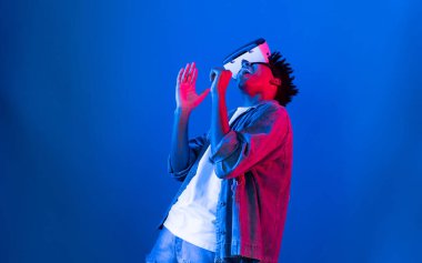 VR gözlükleri kullanarak genç Afrikalı Amerikalı şarkıcı sanal gerçeklik fantezi teknolojisinde mavi ışıklı arka planda karaoke meta uygulama ile şarkı söylemekten zevk alır. Entrika.