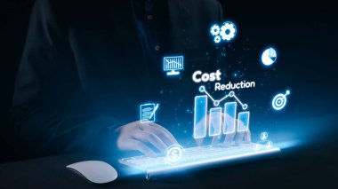 Klavyede oturan profesyonel bir birey, modern bir çalışma ortamında maliyet azaltma ve finansal analiz temalarını gösteren grafikler ve simgelerin holografik görüntüleriyle etkileşim halindedir. Dörtgen