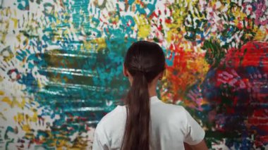 Genç ve güzel bir kızın arka görünüşü renkli duvarı boyamak için elini kaldırır. Çekici bir lise öğrencisi renk paletini tutarken duvarı el iziyle boyuyor. Yaratıcı aktivite. Öğretim