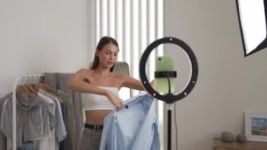Genç sosyal medyum kadın moda videosu yapıyor. Blogcu kameraya selfie çubuğu ve ışık halkası kullanarak gülümserken, aynı zamanda ikna edici çevrimiçi kıyafetlerin izleyicilere veya takipçilere vlog satmasını sağlıyor. Düzenle