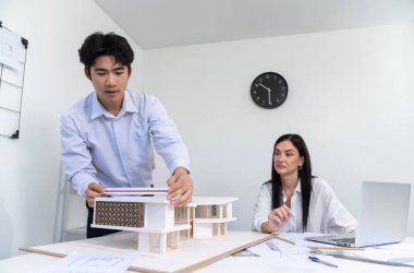 Profesyonel Asyalı erkek mimar ev modelinin uzunluğunu ölçmek için cetvel kullanırken, beyaz tenli genç meslektaşı ev modeliyle toplantı masasındaki verileri analiz etmek için dizüstü bilgisayar kullanıyor. Tertemiz..