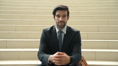 Gülümseyen yetenekli işadamı merdivenlerde otururken kameraya bakıyor. Genç proje yöneticisi, açık havada sarı arka planı bulanık elleri sıkarken kameraya gülümsüyor. Ön manzara. Sevinçli..