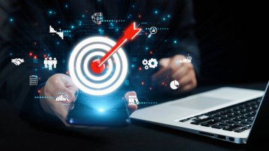 Dijital grafiklerle çevrili, iş stratejisi ve yenilikleri temsil eden parlak bir hedef simgesiyle akıllı bir telefonu tutan resimli bir sahne. Dörtgen