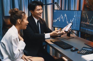 Cep telefonundaki borsa uygulamasını kalemle işaret eden bir ortağa gösteren Asyalı işadamı, gece vakti bilgisayar ekranındaki dijital yatırım veritabanını karşılaştırıyor. İnfobahn.