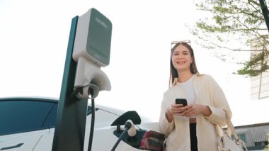 Ağır çekimde genç bir kadın, alışveriş merkezinin otoparkındaki şarj istasyonundan şarj olan EV 'nin pilini şarj ederken akıllı telefondan akü durumunu kontrol ediyor. Modern kadın elektrikli araçla alışverişe gider. Yararlı