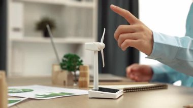 Temiz enerji ve ahşap blok kullanılarak temsil edilen yel değirmeni modelini izleyen iş adamı, gelecekteki yönetim için dizüstü bilgisayar ve defterle çalışan, eko ev dağınıklığını sembolize ediyor. Tanımlama.