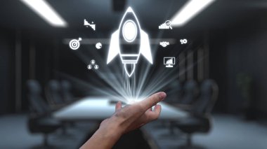 Bir el, yenilik, büyüme ve iş başarısını sembolize eden modern ofis ortamında çeşitli proje yönetim sembolleri arasında parlayan bir roket ikonu yayınlar. Auvana