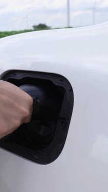 Asyalı adam rüzgar türbiniyle çalışan elektrikli arabaya akıllı EV şarj cihazı takıyor. Sürdürülebilir temiz EV teknolojisi için yenilenebilir enerji kullanımı ve fütüristik dijital batarya durumu hologramı. İnceleyin