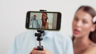 Genç sosyal medyum kadın moda videosu yapıyor. Blogcu kameraya selfie çubuğu ve ışık halkası kullanarak gülümserken, aynı zamanda ikna edici çevrimiçi kıyafetlerin izleyicilere veya takipçilere vlog satmasını sağlıyor. Düzenle