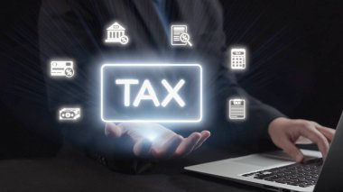 Bir iş adamı, vergilendirmenin modern finansmandaki önemini temsil eden yüzen finansal simgelerle çevrili bir dizüstü bilgisayarın üzerinde parlak bir vergi tabelası tutuyor. Dörtgen