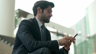 Gülümseyen yetenekli işadamı merdivenlerde otururken kameraya bakıyor. Genç profesyonel proje yöneticisi arka planda bulanık cep telefonunu tutarken kameraya gülümsüyor. Sevinçli..