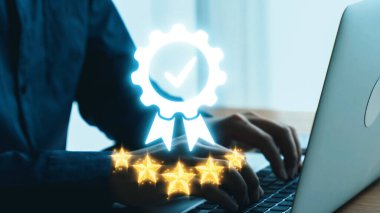 Dijital rozet ve yıldızlarla tamamlanmış, modern ve profesyonel bir ortamda kalite ve başarıyı simgeleyen bir dizüstü bilgisayarda yazı yazan ellerin yakın çekimi. Dörtgen