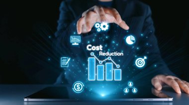 Bir işadamı, modern bir çalışma alanındaki finans ve teknolojik yenilikleri vurgulayarak, bar grafikleri ve simgeler kullanarak dijital bir maliyet azaltma kavramını resmetmektedir. Dörtgen