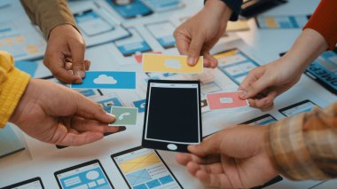 Değişik bireylerden oluşan bir grubun mobil bir uygulama tasarladığı dinamik bir sahne. Eller, tellerle çevriliyken renkli simgeleri ve ekranları tutar. SACTR