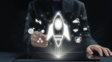 Bir iş adamı, modern çalışma alanındaki büyüme, teknoloji ve yaratıcılığı vurgulayan yenilikçi projelerin başlatılmasını gösteren bir roket ikonunu görselleştirmek için bir tablet kullanır. Dörtgen