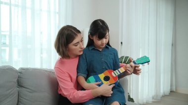 Kafkasyalı bir anne evde akustik müzik öğretirken ukulele çalan mutlu kız. Sevimli çocuk enstrüman öğreniyor. Kafkasyalı mutlu anne ve kız birlikte vakit geçiriyorlar. Pedagoji.